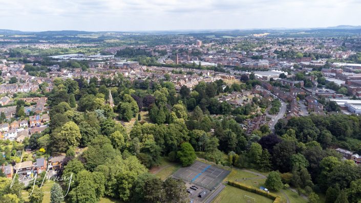 Aerial view of a green park with trees and tennis courts, surrounded by houses and roads, with a sprawling town and rolling hills visible in the background under a clear sky.