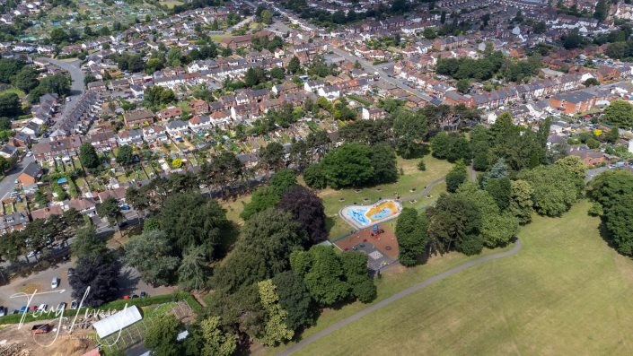 Aerial view of a residential neighbourhood with rows of houses, tree-lined streets, and a large park featuring a playground with a blue and yellow play area, surrounded by green lawns and dense trees.
