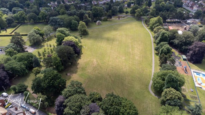 Aerial view of a large grassy park surrounded by trees, with walking paths, a playground, scattered benches, and houses along the edges. The park is lush and green under daylight.