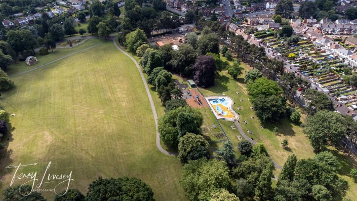 Aerial view of a large park with open grassy areas, a winding path, a colourful playground, and clusters of trees, bordered by rows of houses and gardens.