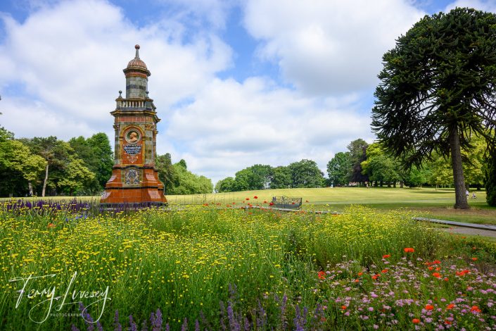 A Victorian clock tower stands amid vibrant wildflowers in a park under a partly cloudy blue sky, bordered by green trees and a well-kept lawn.