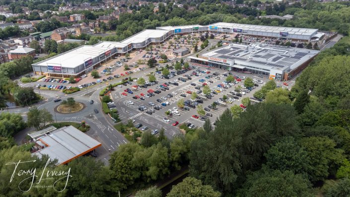 Aerial view of a large retail park with multiple shops, a main car park filled with cars, surrounded by trees and residential houses in the background. A road and smaller building are visible in the foreground.