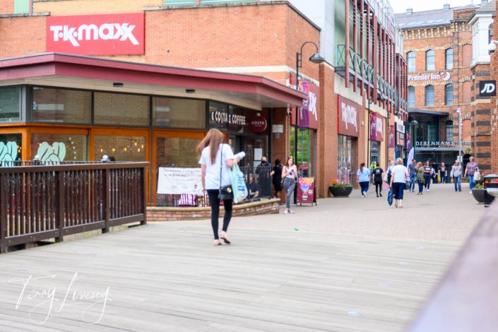 A busy shopping street with people walking past shops like TK Maxx, Costa Coffee, JD, and Premier Inn on a cloudy day. Some pedestrians are carrying shopping bags.