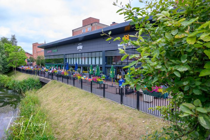 A modern riverside café with outdoor seating, colourful flower planters, and large windows. People are dining outside, and lush greenery surrounds the area. A black fence runs along the water's edge.