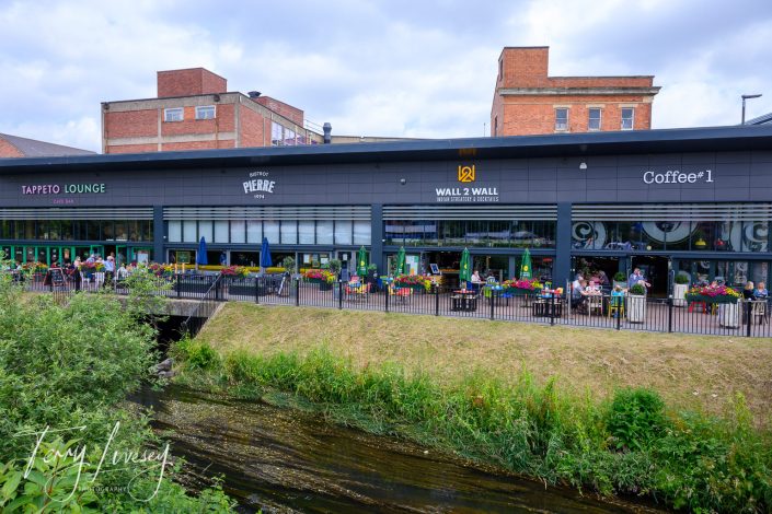 Modern building with several cafés and restaurants facing a canal, including Tappeto Lounge, Bistrot Pierre, Wall 2 Wall, and Coffee#1. People are sitting at outdoor tables, and there are colourful flowers and greenery.