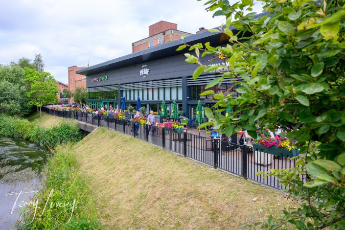 A modern riverside restaurant with a black exterior, large windows, and colourful flowers on the outdoor terrace. People are seated at tables along the water, enjoying the greenery and pleasant atmosphere.