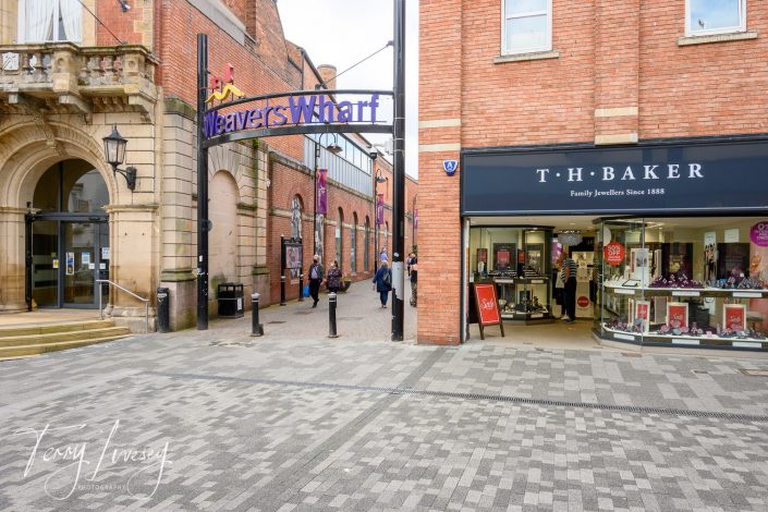 A pedestrian shopping area with the sign Weavers Wharf overhead, people walking, and the shopfront of T.H. Baker jewellers on the right. The scene features brick buildings and a paved walkway.