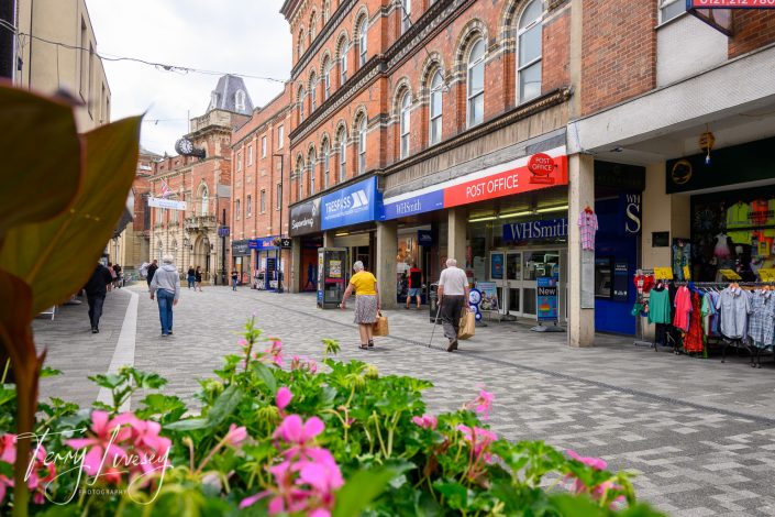 A pedestrianised street with people walking past shops, including WHSmith and the Post Office. Pink flowers are in the foreground, and historic brick buildings line the street under a partly cloudy sky.