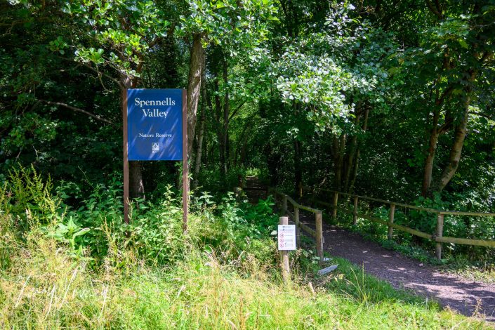 A blue sign reads Spennells Valley Nature Reserve at the entrance to a wooded path, surrounded by lush green foliage and a wooden railing. Sunlight filters through the trees onto the footpath.