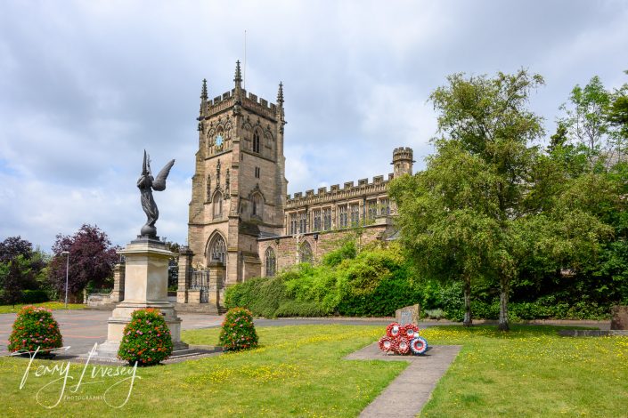 An historic stone church with a tall clock tower stands behind a war memorial statue of an angel and a display of red and white wreaths on a green lawn, surrounded by trees and cloudy skies.