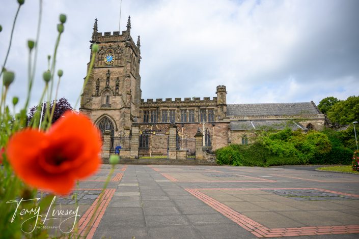 A historic stone church with a clock tower stands in the background, while bright red poppies and green plants are in the foreground. The sky is partly cloudy, and paved paths lead up to the building.
