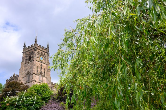 A stone church tower with a clock rises above green trees and leafy branches against a partly cloudy sky.