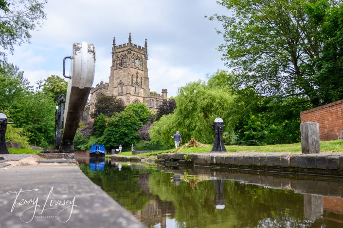A peaceful canal scene with a narrowboat approaching a lock gate, lush greenery on both sides, and a historic stone church with a clock tower in the background under a cloudy sky.