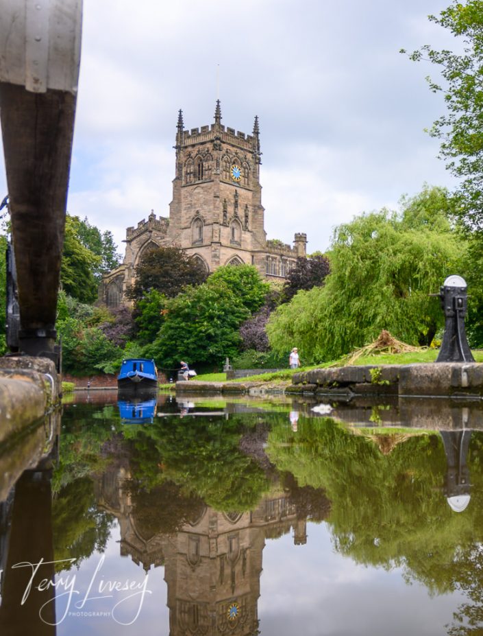 A stone church with a clock tower is reflected in a calm canal, surrounded by lush greenery. A blue boat floats nearby, and a person is visible on the path beside the water.