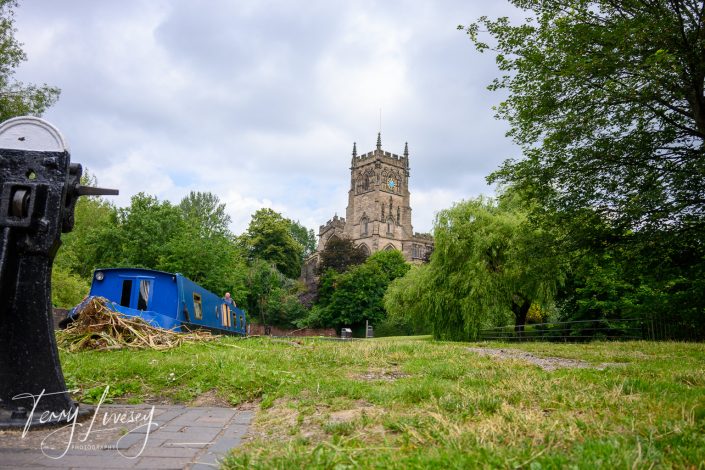 A blue canal boat is moored on a grassy bank near a tree-lined path, with a historic stone church and clock tower rising in the background under a partly cloudy sky.
