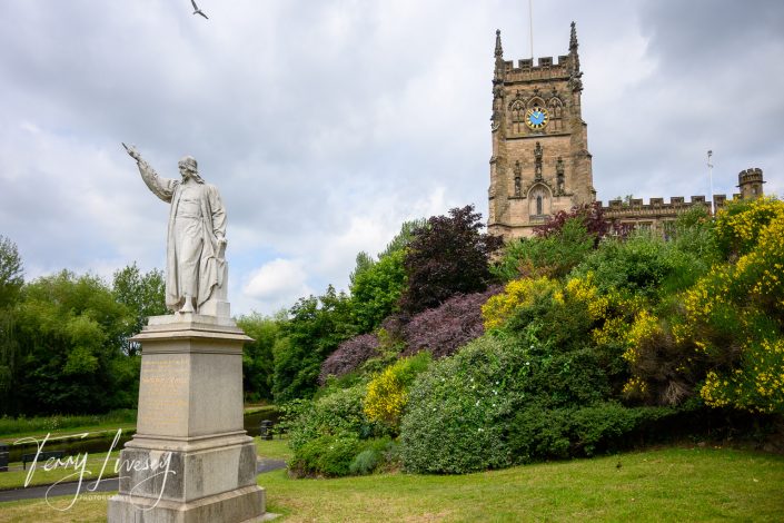 A white statue of a person stands in front of lush greenery and a historic stone church tower with a clock. Colourful bushes and trees surround the area under a partly cloudy sky.