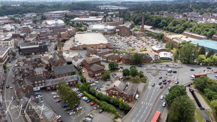 Aerial view of a town with a mix of historic and modern buildings, car parks, roads with cars, green trees, and a large park area in the background under a cloudy sky.
