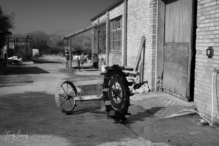 A black and white photo of a rustic farmyard shows an old, weathered brick building with large wooden doors. A metal, antique farming machine with large wheels is positioned in the foreground on a concrete surface.