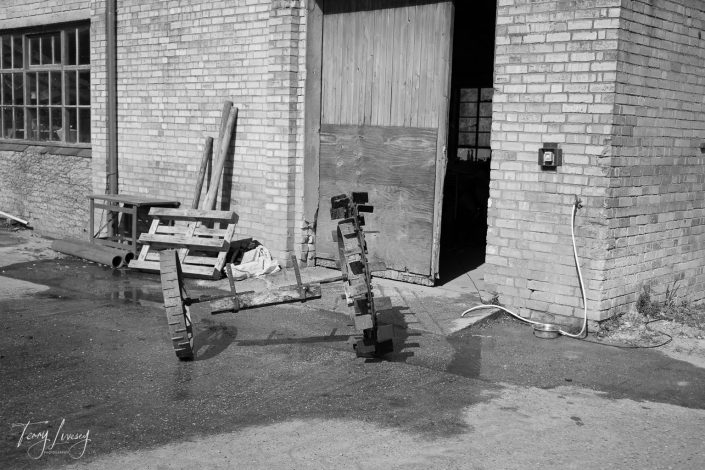 Two abstract, animal-like sculptures made of stacked bricks stand on a wet concrete surface outside a brick building with wooden doors and windows. Sunlight casts shadows; a hose and pallets are visible nearby.