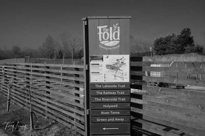 A wooden sign for The Fold lists six walking trails, including The Lakeside Trail and The Railway Trail. The background features wooden fencing, trees, and clear sky. The photo is in black and white.