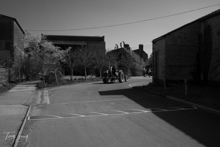 A black and white photo of a quiet rural street, with a person driving a vintage tractor away from the camera. Trees, old farm buildings, and houses line the road under a clear sky.