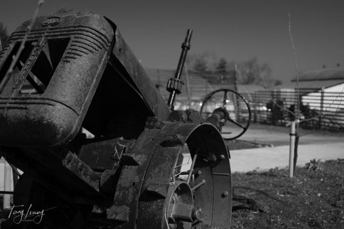 Black and white photo of a rusty, vintage tractor in the foreground, partially seen from the side, with farm buildings, fencing, and bare trees blurred in the background.