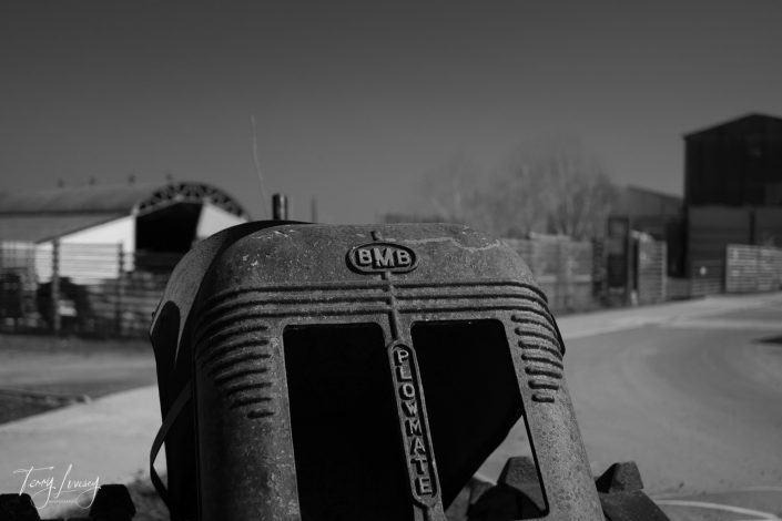 A close-up, black-and-white photo of the front of an old BMB Plowmate tractor, with blurred sheds and fencing in the background and a clear sky above.