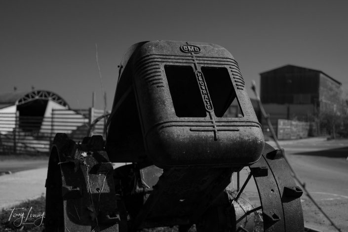 Black and white photo of an old, rusted BMB Plowmate tractor seen from the front, with farm buildings and a road in the blurred background. The scene evokes a sense of rural decay and history.