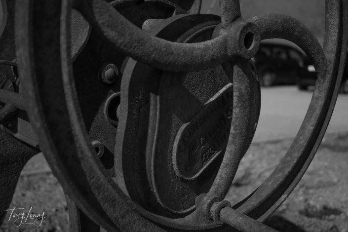 Close-up of a large, weathered iron wheel with industrial machinery details, captured in black and white. A blurred car and road are visible in the background.
