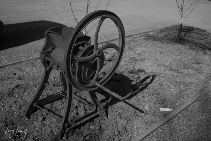 An old, rusted metal machine with a large wheel sits on a dirt patch under sunlight. Sparse tree saplings are planted nearby, and a faint shadow of the machine falls on the ground.
