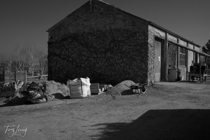 A black and white photo of a large shed with climbing vines, piles of gravel, large sacks, a wheelbarrow, and scattered building materials outside on a sunny day. Shadows stretch across the ground.