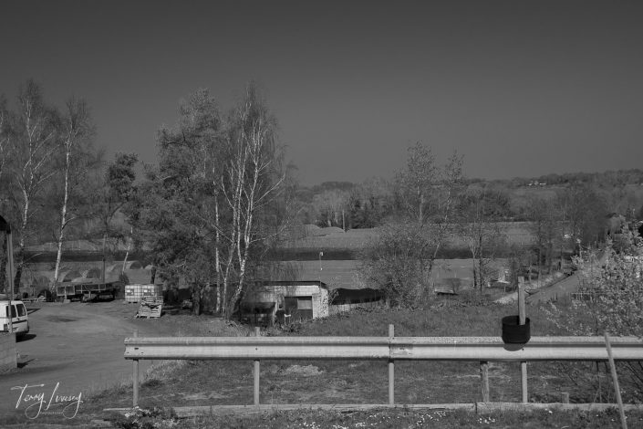 Black and white photo of a rural landscape with trees, a small road, and a metal crash barrier in the foreground. There are parked vehicles, a river or pond, and distant rolling hills under a clear sky.