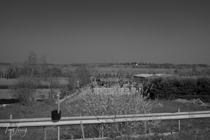Black and white photo of a rural landscape with fields, scattered trees, farm buildings, and stacks of materials. In the foreground, a potted plant sits on a wooden fence. The horizon shows a tree-lined ridge.