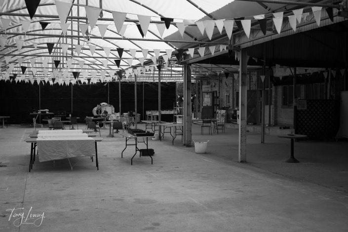 A black and white photo of an empty outdoor event space with tables, chairs, and flags hanging from the ceiling. The area is covered by a partial roof and appears to be set up for a gathering or market.