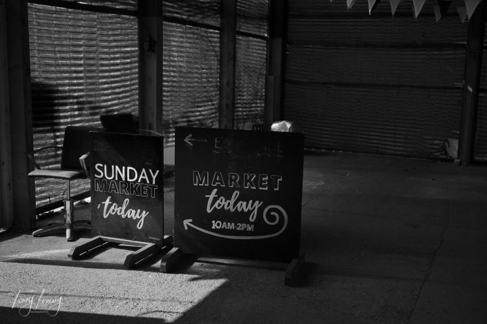 Two black signs in a shaded area read SUNDAY MARKET today and MARKET today 10AM–2PM with arrows pointing left. Empty chairs and bamboo walls are visible in the background. Black and white photo.