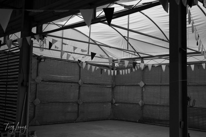 A black and white photo of an empty indoor space with a tented roof, strung triangular bunting hanging across the area, and corrugated metal walls. The scene appears quiet and unused.