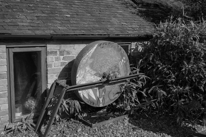 A large, weathered round metal object is mounted on a metal frame outside a brick building with a tiled roof. Tall plants grow nearby, and a window is visible to the left. The scene is in black and white.