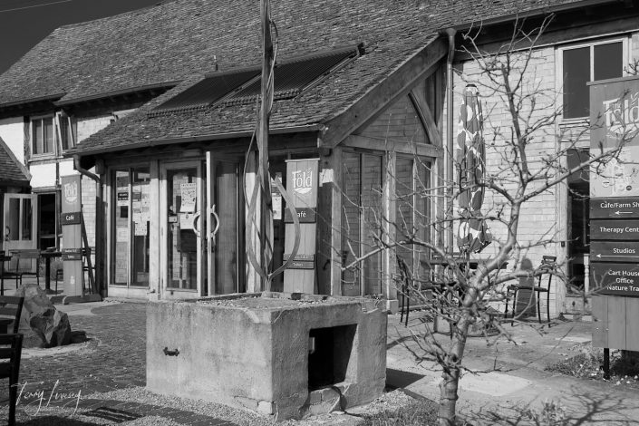 A black and white photo of a rustic building with wooden beams and a tiled roof, housing a café and therapy centre called The Fold. A leafless tree and a stone planter are visible in the courtyard.