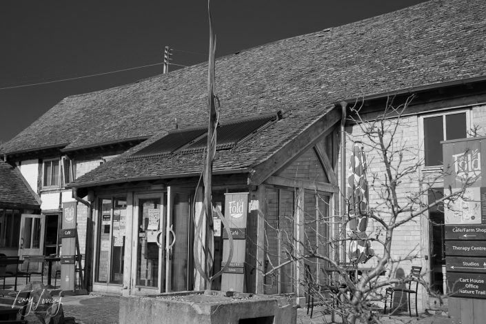 A rustic building with a tiled roof and glass entrance, featuring banners for a café and heritage centre. Bare tree branches are visible in the foreground under a clear sky.