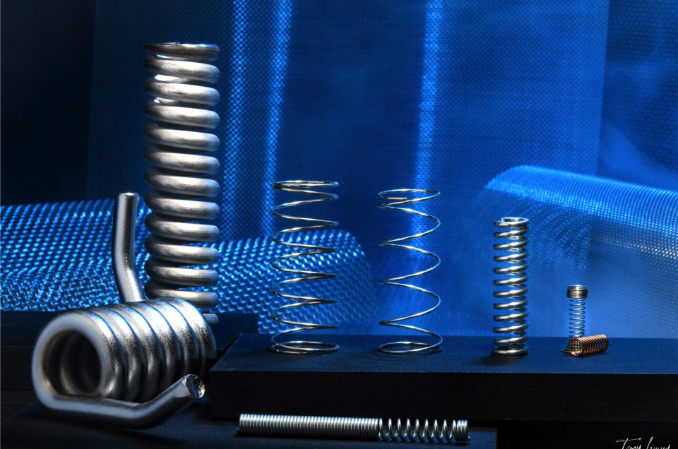 A variety of metal springs in different shapes and sizes are arranged upright and lying flat on black surfaces, with a blue mesh fabric backdrop. The lighting highlights the metallic texture of the springs.