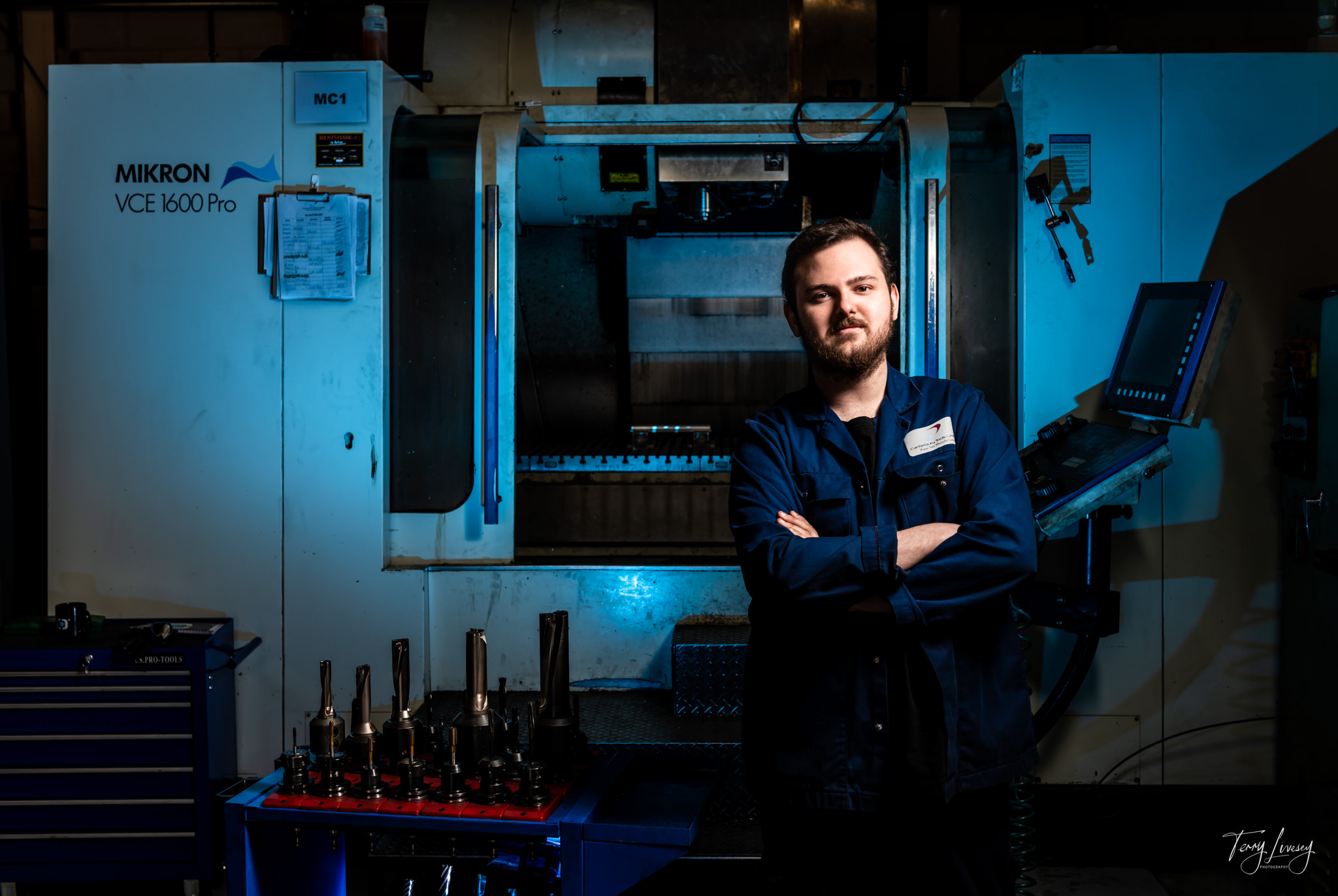 A man in a blue work uniform stands confidently with arms folded in front of a large industrial CNC machine labelled Mikron VCE 1600 Pro, captured in striking photography with various machine tools displayed on a nearby table.