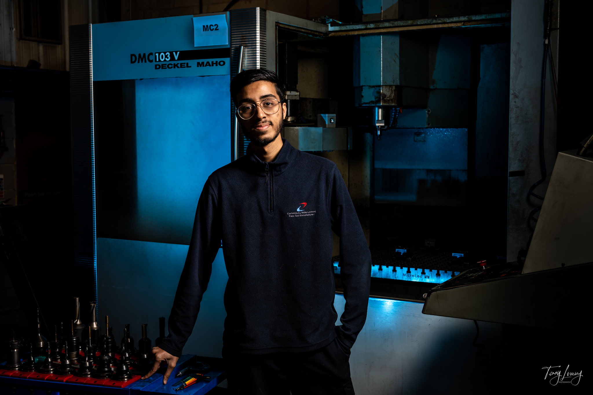 A young man wearing glasses and a dark jumper stands confidently in front of industrial machinery in a dimly lit workshop, captured through striking photography, with various metal tools arranged on a blue table beside him.