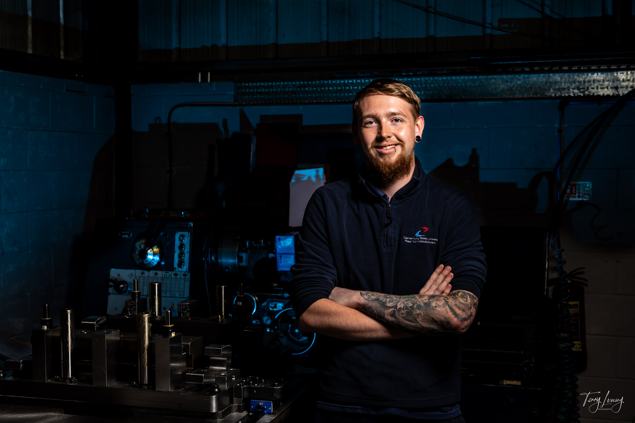 A man with a beard and tattoos stands smiling with arms crossed in a dimly lit industrial workshop, machinery and metal parts visible in the background, capturing the essence of industrial photography.