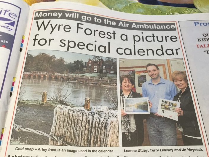 A newspaper article features a headline about Wyre Forest photography for a charity calendar. The image shows three people holding the calendar and a smaller photo of a frosty riverside scene.