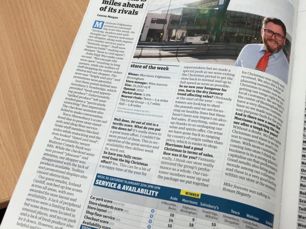 An open magazine on a wooden table features an article about Morrisons, including impressive photography of a Morrisons store and a smiling man in a shirt and tie outside. Text and comparison tables are also visible.