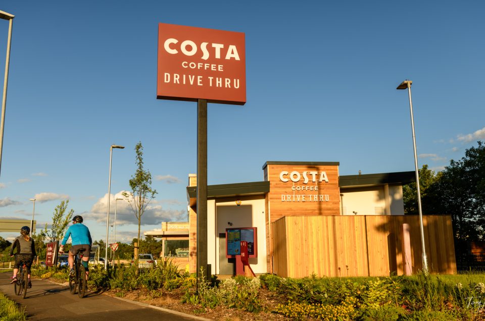 Two people ride bicycles past a Costa Coffee drive-through on a sunny day. The modern building with wood panelling and the bold red signage creates a perfect scene for photography enthusiasts capturing city life moments.