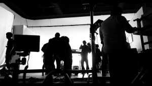 Silhouetted crew members work on a photography set filled with equipment, lights, and monitors, against a bright white backdrop in a studio.