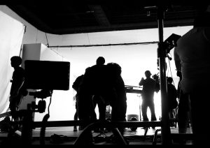 Silhouettes of several people working on a photography or film shoot set, surrounded by equipment, lights, and cables in a large studio with a white backdrop.