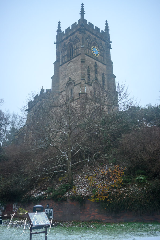 A historic stone church tower with clock face stands surrounded by trees and an informational sign in the foreground.