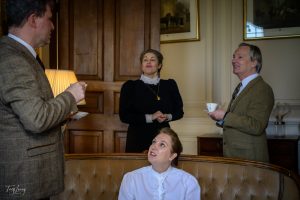 Four people in vintage clothing talk and drink tea in an elegant room, captured in a timeless photography style. One woman sits on a sofa looking up whilst the others stand nearby amid framed art and wood panelling.